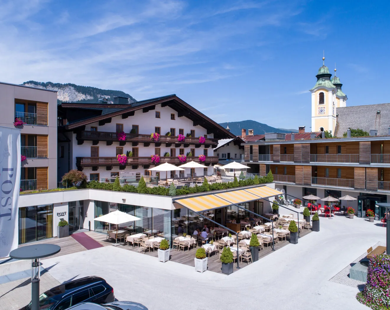 Blick auf ein Hotel mit Terrasse, Tischen und Stühlen, Bergen im Hintergrund.