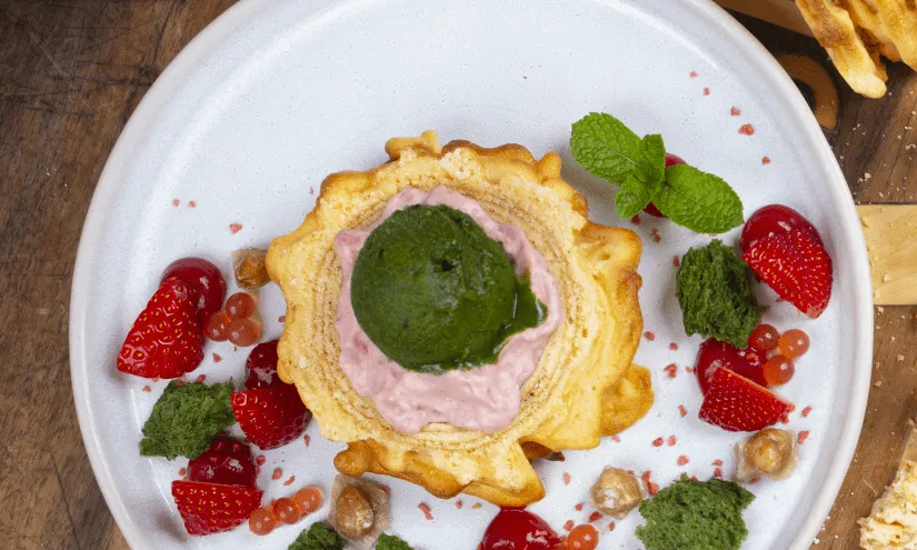Dessert mit grüner und rosa Mousse, Erdbeeren, Nüssen und Kräutern auf einem Teller