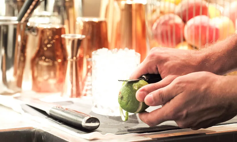 Eine Hand schneidet eine Limette auf einer Theke mit verschiedenen Barwerkzeugen und Früchten im Hintergrund