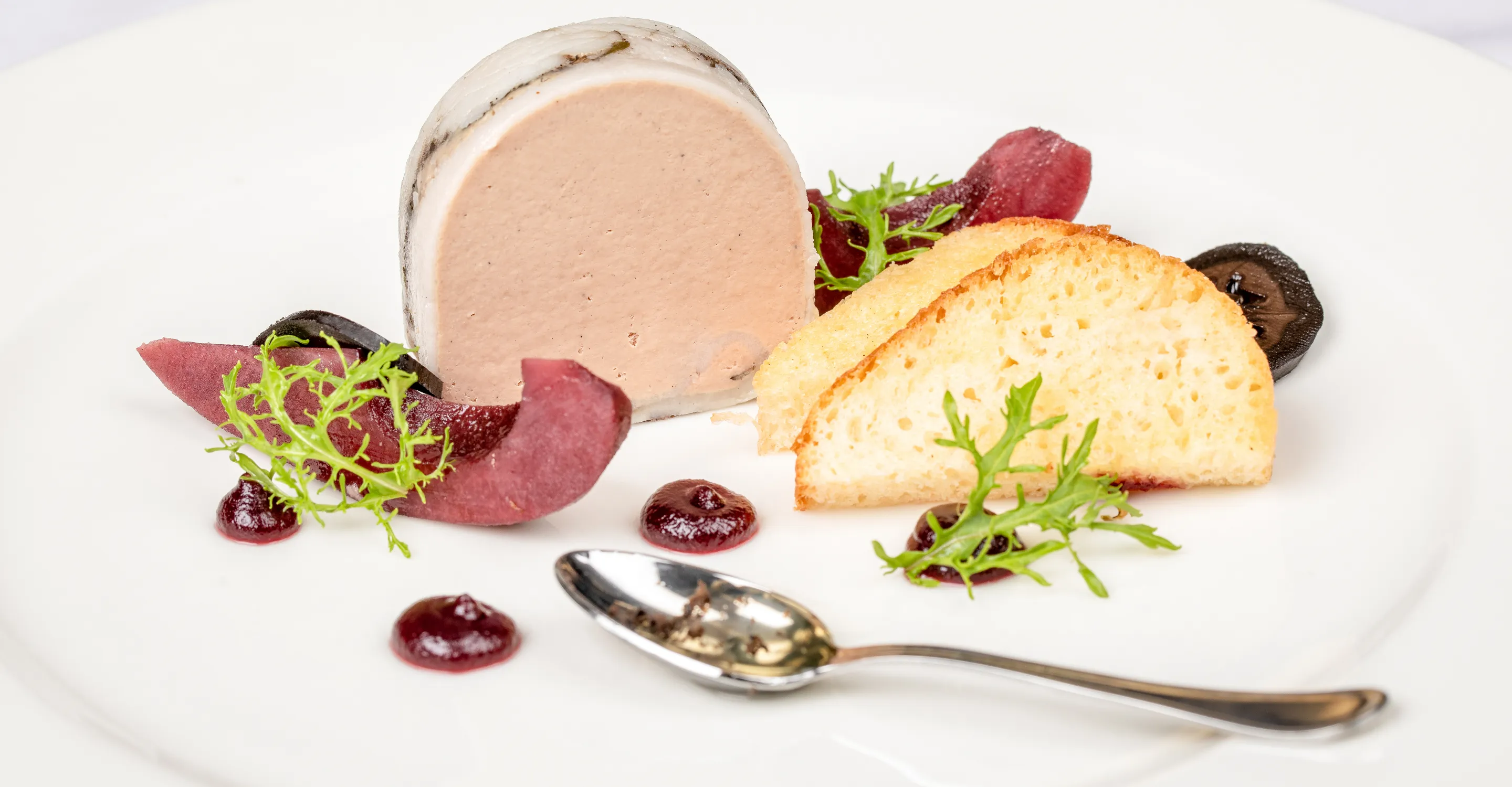 Gericht mit einem Stück Mousse, Brot, Beeren und Kräutern auf einem Teller