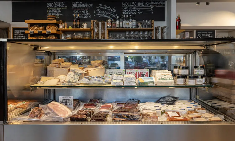Kühlschrank mit verschiedenen Fleisch- und Käseprodukten, im Hintergrund ein Regal mit Gläsern